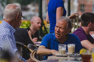 Zwei Männer diskutieren miteinander an einem Tisch mit zwei Kaffees darauf. Sie sitzen auf der Trasse von einem Café